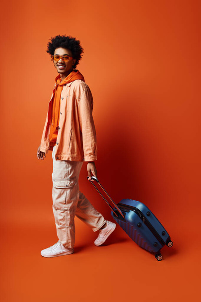 Модний афроамериканський чоловік з кучерявим афро волоссям, що тримає синю валізу на помаранчевому тлі, виглядає емоційно. - Фото, зображення