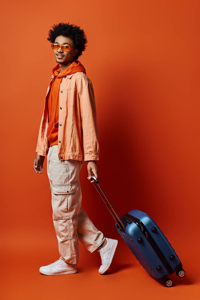 オレンジ色の背景に青いスーツケースを運ぶオレンジ色のジャケットのカーリーヘアを持つファッショナブルなアフリカ系アメリカ人男性. - 写真・画像