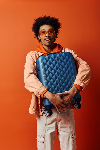 Homme afro-américain élégant en tenue tendance et lunettes de soleil tenant un bagage bleu sur un fond orange. - Photo, image