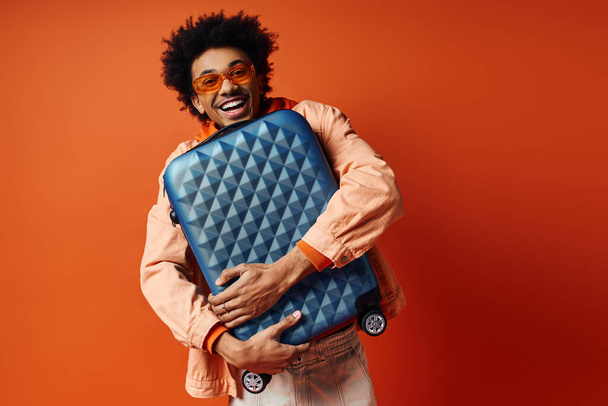 Stylowy młody Afroamerykanin z kręconymi włosami i modnym strojem stoi na pomarańczowym tle, trzymając niebieską część bagażu. - Zdjęcie, obraz