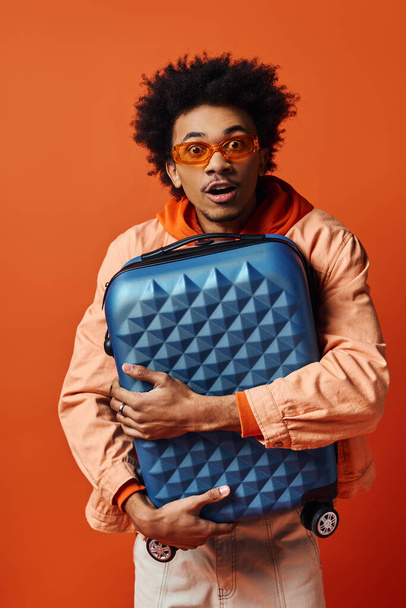 Egy elegáns, fiatal afro-amerikai férfi göndör hajjal, kezében egy kék bőrönddel, narancssárga háttérrel.. - Fotó, kép