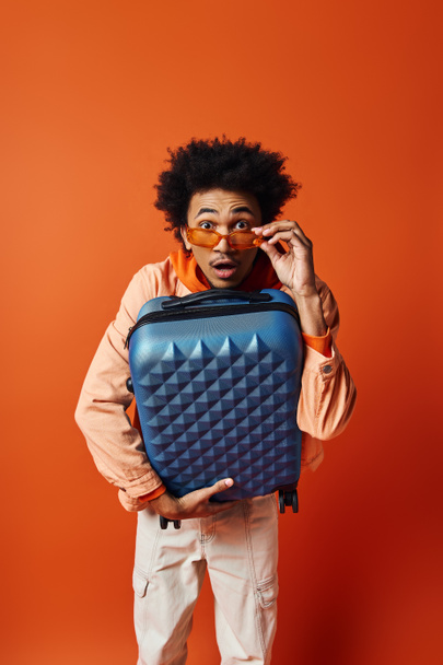 Homme afro-américain tendance aux cheveux bouclés tenant un élégant bagage bleu sur un fond orange. - Photo, image
