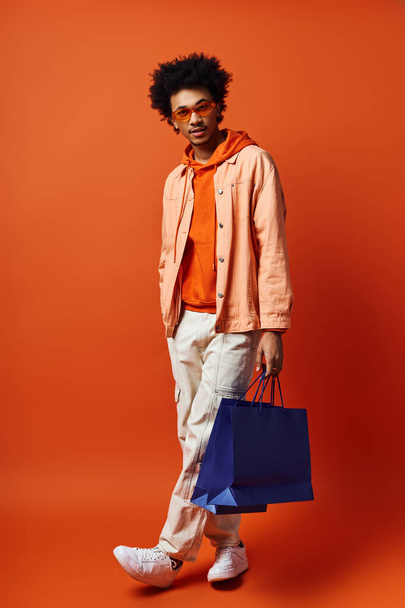 Młody Afroamerykanin w pomarańczowej koszuli i białych spodniach, trzymający niebieską torbę, na jasnym pomarańczowym tle. - Zdjęcie, obraz