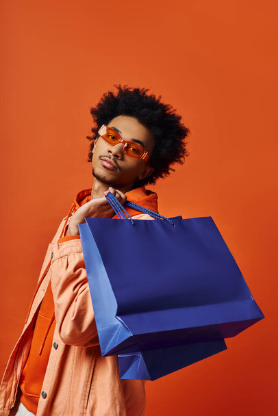 Кудрявый афроамериканец в модном наряде держит синюю сумку для покупок на оранжевом фоне, выглядящую эмоционально. - Фото, изображение