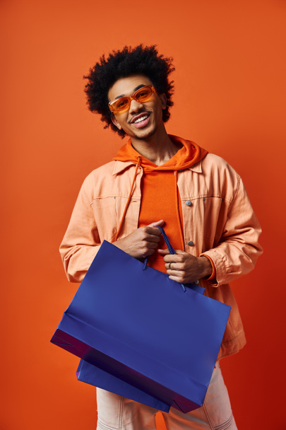 オレンジ色の背景に青いショッピングバッグを持っているサングラスのスタイリッシュな若いアフリカ系アメリカ人男性. - 写真・画像