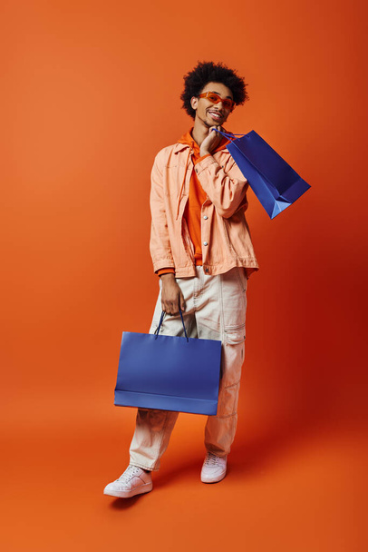 Homme afro-américain bouclé en tenue tendance tient un sac bleu tout en mettant en valeur un sac bleu unique et vibrant sur un fond orange. - Photo, image