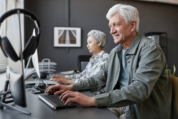 Πλάγια άποψη πορτρέτο του χαμογελαστού ηλικιωμένου άνδρα που χρησιμοποιεί υπολογιστή στην τάξη για ηλικιωμένους, αντιγραφή χώρου - Φωτογραφία, εικόνα