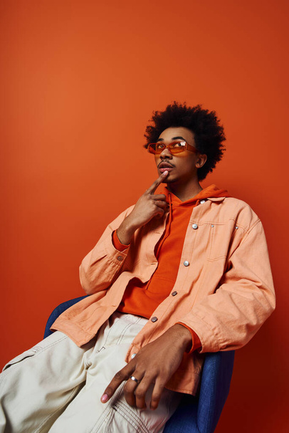 Egy elegáns, fiatal afro-amerikai férfi, göndör hajjal, kék székben ülve, narancssárga ingben, gondolataiba merülve.. - Fotó, kép