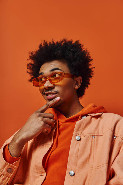 Ein trendiger junger afroamerikanischer Mann mit lockigem Haar, orangefarbenem Hemd und Sonnenbrille posiert vor orangefarbenem Hintergrund. - Foto, Bild