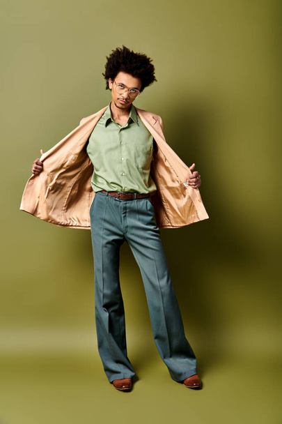 Un giovane uomo afroamericano elegante con i capelli ricci in piedi con fiducia di fronte a un muro verde audace mentre indossa abbigliamento alla moda e occhiali da sole. - Foto, immagini