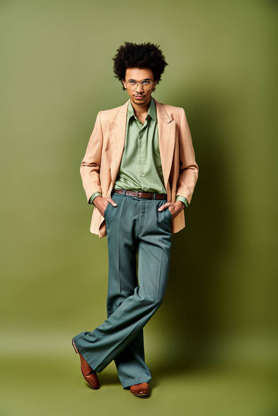 Un jeune homme afro-américain élégant aux cheveux bouclés se tient avec confiance devant un mur vert vibrant, portant des vêtements à la mode et des lunettes de soleil. - Photo, image