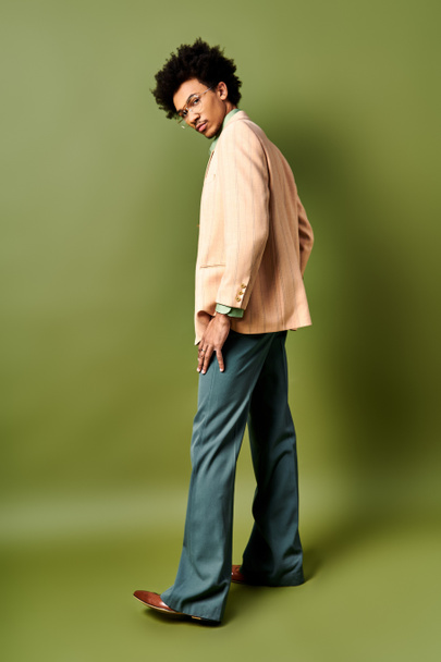 Un hombre afroamericano de moda con el pelo rizado y gafas de sol de pie con confianza delante de una pared verde vibrante. - Foto, imagen