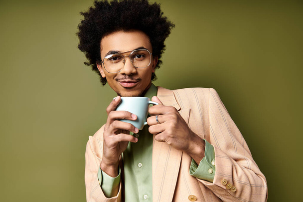 Ένας μοντέρνος νεαρός Αφροαμερικανός με σγουρά μαλλιά και γυαλιά ηλίου κρατά ένα φλιτζάνι καφέ στον ατμό σε ένα πράσινο φόντο. - Φωτογραφία, εικόνα