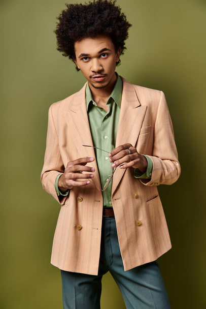 Стильный молодой афроамериканец с вьющимися волосами, в коричневой куртке и зеленой рубашке, в модном наряде и солнечных очках. - Фото, изображение
