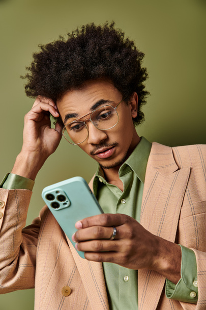 Κομψός νεαρός Αφροαμερικάνος με κοστούμι και γυαλιά ηλίου που κρατά με αυτοπεποίθηση ένα κινητό τηλέφωνο σε πράσινο φόντο. - Φωτογραφία, εικόνα