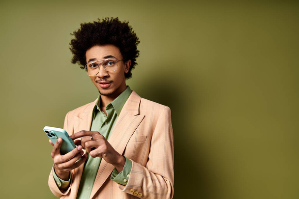 Ένας κομψός νεαρός Αφροαμερικανός με κοστούμι και γυαλιά ηλίου, απορροφημένος στο κινητό του σε πράσινο φόντο.. - Φωτογραφία, εικόνα