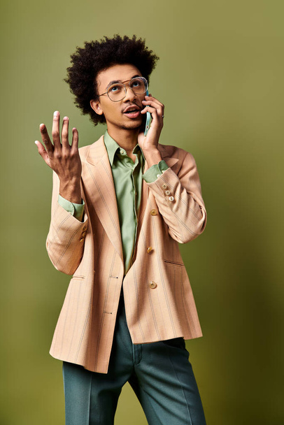 Tyylikäs nuori afroamerikkalainen mies puvussa ja aurinkolasit puhumassa matkapuhelimella elinvoimaista vihreää taustaa vasten. - Valokuva, kuva