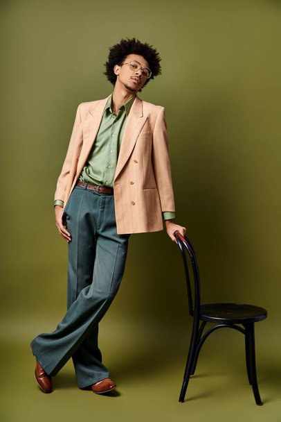 Trendikäs, kiharatukkainen afroamerikkalainen mies tyylikkäässä puvussa seisoo tuolin vieressä elinvoimaista vihreää seinää vasten.. - Valokuva, kuva