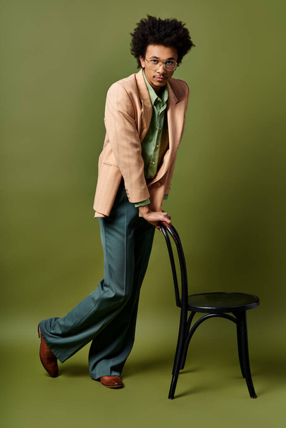 Un hombre afroamericano elegante y rizado se apoya pensativamente en una silla, usando atuendos de moda y gafas de sol contra un fondo verde.. - Foto, imagen