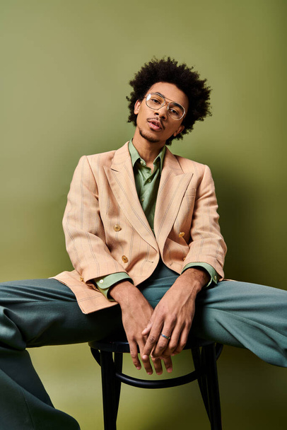 Un giovane afroamericano elegante con i capelli ricci seduto su una sedia, gambe incrociate, in abbigliamento alla moda e occhiali da sole su uno sfondo verde. - Foto, immagini