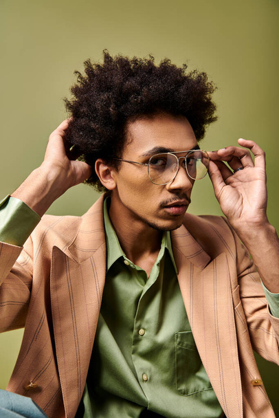 Κομψός νεαρός Αφροαμερικανός με σγουρά μαλλιά, κοφτερό κοστούμι και γυαλιά ηλίου, κάθεται σε πράσινο φόντο. - Φωτογραφία, εικόνα