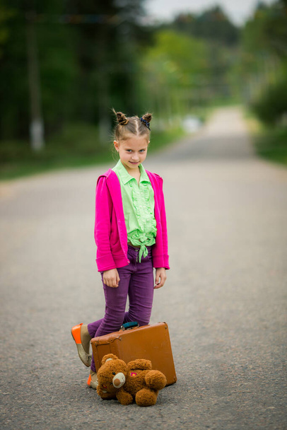 Στην άκρη του δρόμου, ένα κορίτσι στολισμένο με φωτεινά ρούχα κουβαλάει τη βαλίτσα της και το αγαπημένο της βελούδινο παιχνίδι, συμβολίζοντας τον ενθουσιασμό της ανακάλυψης.. - Φωτογραφία, εικόνα