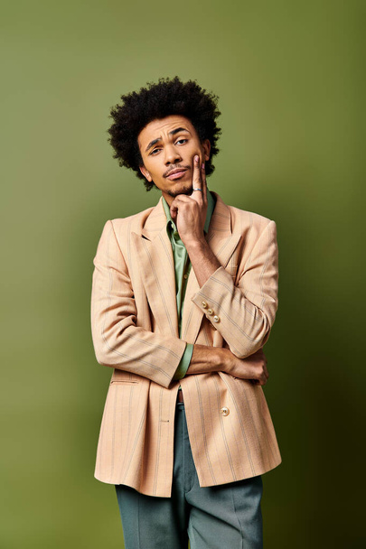 Молодой афроамериканец в модном наряде стоит с руками на подбородке в задумчивой позе на зеленом фоне. - Фото, изображение