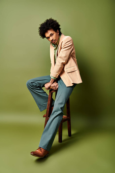 Een stijlvolle jonge Afro-Amerikaanse man in trendy kleding zittend op een houten stoel tegen een groene achtergrond. - Foto, afbeelding