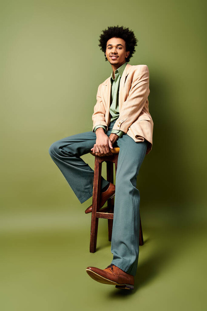 Un jeune homme afro-américain élégant aux cheveux bouclés s'assoit en toute confiance sur un tabouret en bois, vêtu d'une tenue tendance - Photo, image
