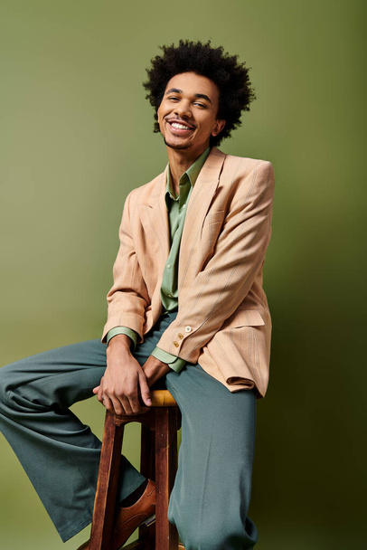 Kıvırcık saçlı şık bir Afrikalı genç Amerikalı, ahşap bir taburede oturuyor, yeşil arka planda moda kıyafetler giyiyor.. - Fotoğraf, Görsel