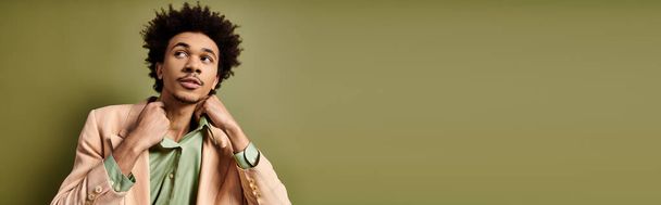 A stylish, young African American man with curly hair leans against a green wall, exuding confidence in trendy attire - Photo, Image