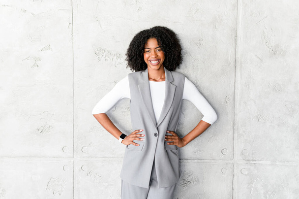 A poised African-American businesswoman stands with hands on her hips, wearing a stylish grey sleeveless vest and trousers, radiating confidence and professionalism against a textured grey background. - Foto, Imagem