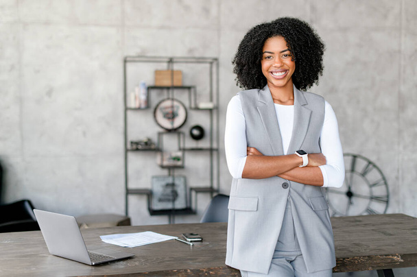 A beaming African-American woman stands confidently, her arms crossed, in a stylish business-casual vest and white long-sleeve shirt, representing a blend of modern professionalism and approachability - 写真・画像