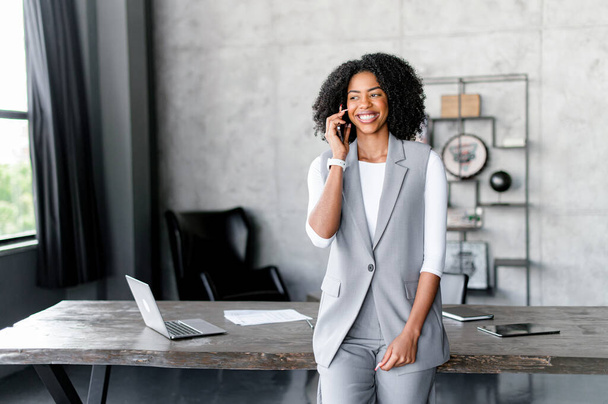 A joyful African-American businesswoman engages in a phone call, her expression one of satisfaction and command, set against the backdrop of an industrial-chic office space - Foto, imagen