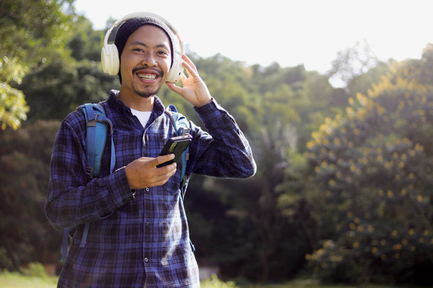 Junger asiatischer Mann in lässigem Outfit, mit Mütze, kariertem Hemd, Rucksack und Kopfhörer, blättert in seinem Handy nach Musik und sucht scheinbar die perfekte Playlist oder den perfekten Song für seinen morgendlichen Spaziergang in der Natur aus.. - Foto, Bild
