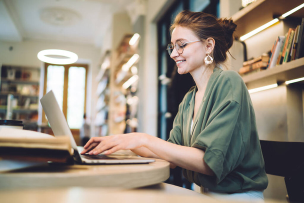 Side view of smiling adult modern female in casual outfit and with glasses sitting at table with books typing laptop against blurred shelves in modern library - Fotografie, Obrázek
