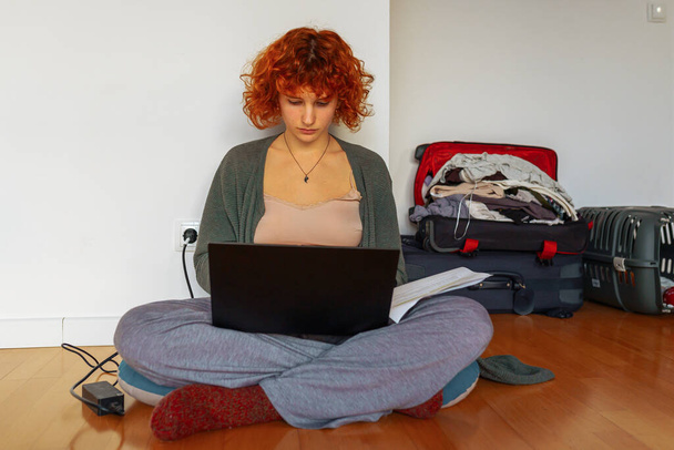 Portret rudowłosej nastolatki siedzącej na podłodze w pustym, nieumeblowanym pokoju, korzystającej z tabletu, niezmontowanych walizek stojących w pobliżu, przenoszącej koncepcję, wynajmującej mieszkania dla studentów, studiującej online w domu - Zdjęcie, obraz