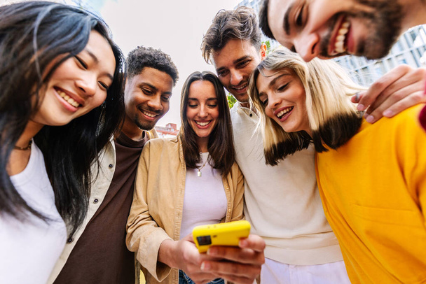 Skupina mladých šťastných lidí, kteří používají mobilní telefon na City Street. Millennial rozmanité přátele těší obsah sociálních médií v mobilní aplikaci. Technologický životní styl a koncepce společenství mládeže. - Fotografie, Obrázek