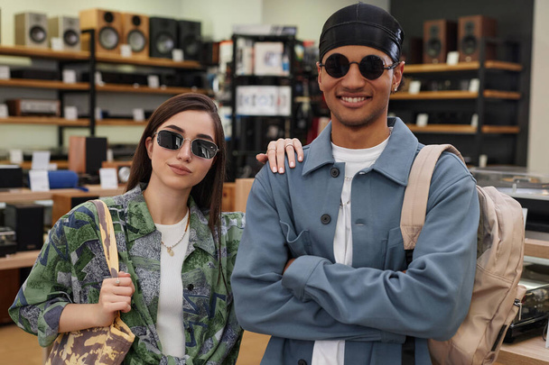 Cintura hacia arriba retrato de la pareja joven funky usando gafas de sol mientras está de pie en la tienda de música y mirando a la cámara - Foto, imagen