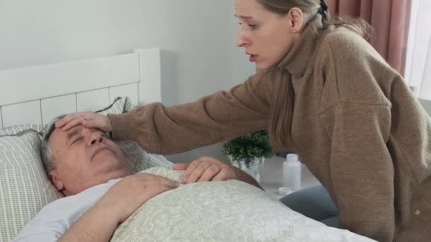 Anxious adult daughter touches forehead of her sick elderly father and feel feever. Scared woman sits on a bed with ill senior man. Family care concept. - Footage, Video