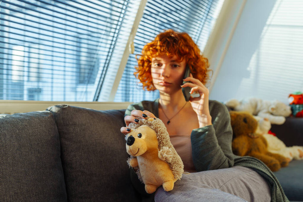 muotokuva punatukkainen kihara teini tyttö käyttää puhelinta istuessaan mukava sohva kotona, lähellä suuri ikkuna kaihtimet. halauksia lasten leluja, nostalgisia tunteita. varjot kaihtimet putoavat kasvot - Valokuva, kuva