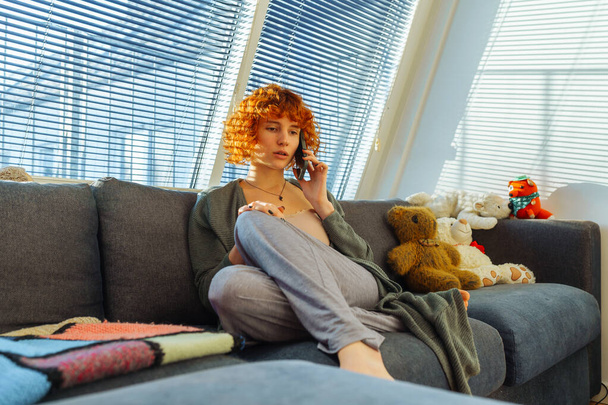 Portret rudowłosy kręcone nastolatka za pomocą telefonu podczas siedzenia na wygodnej kanapie w domu, w pobliżu dużych okien z żaluzjami. nostalgiczne emocje. Cienie z żaluzji padają na twarz. wiele tła zabawki - Zdjęcie, obraz