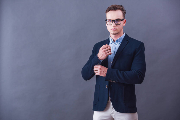 Knappe jonge zakenman in pak en brillen is manchetknopen aanpassen en kijken naar camera, op grijze achtergrond - Foto, afbeelding