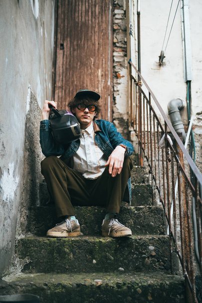 Hipper junger Mann im Vintage-Look sitzt mit Retro-Kassettenspieler auf Steinstufen in urbaner Umgebung und strahlt Sinn für Retro-Mode und Kontemplation aus.  - Foto, Bild