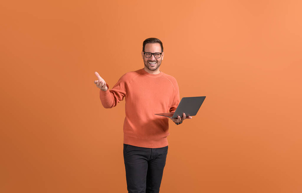Przystojny przedsiębiorca trzymając laptopa i gestykulując pewnie stojąc na pomarańczowym tle - Zdjęcie, obraz