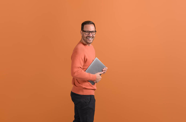 Портрет веселого молодого человека с ноутбуком и смеющегося над камерой на изолированном оранжевом фоне - Фото, изображение