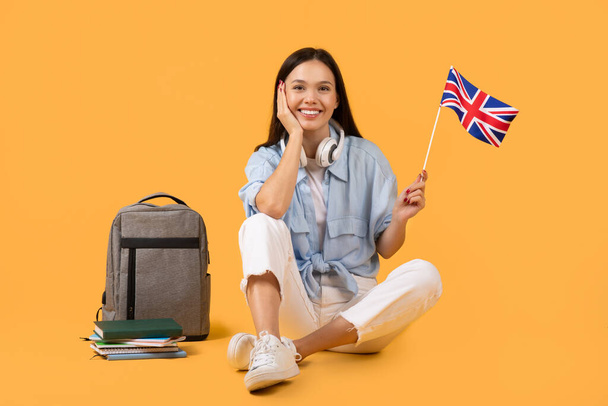 Young woman learner with headphones holding a United Kingdom flag, sitting next to a backpack and books - Photo, Image