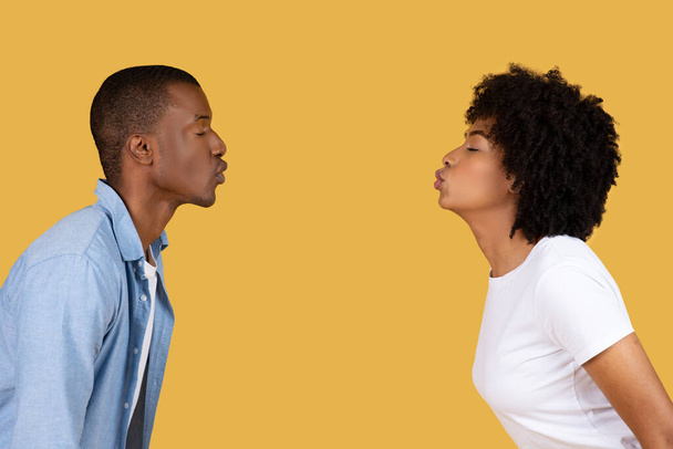 Мужчина и женщина стоят лицом друг к другу в профиль с простым желтым фоном, афроамериканская пара целуется - Фото, изображение