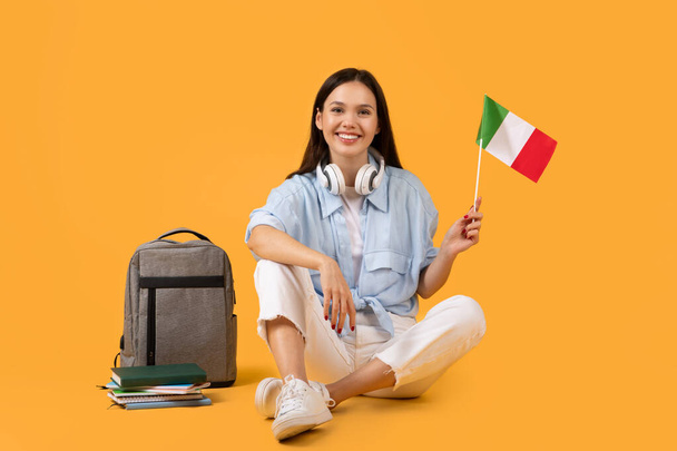 Cheerful young woman showcasing an Italian flag, accompanied by headphones and educational material - Photo, Image