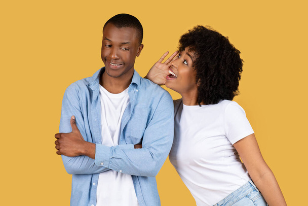 Eine junge Afroamerikanerin flüstert einem Mann ins Ohr, dass sie ein Geheimnis auf gelbem Hintergrund teilen. - Foto, Bild